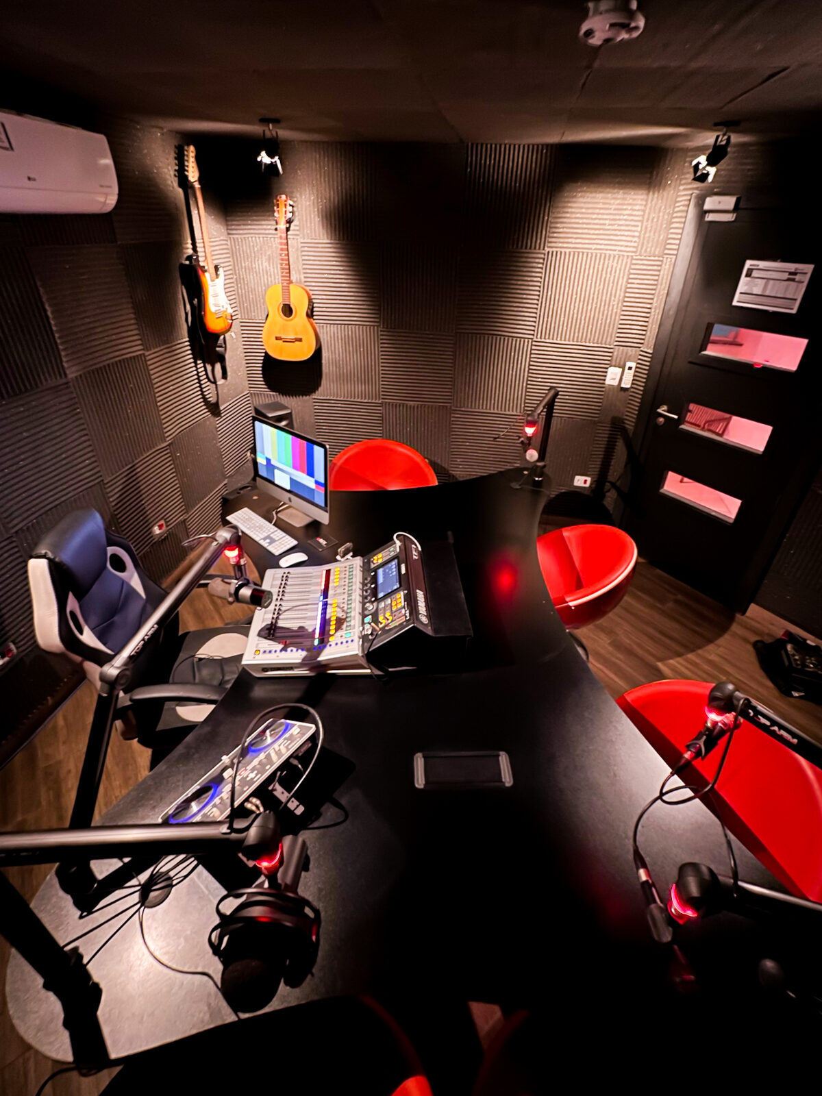 Estúdio A visto de cima: mesa de podcast com microfones em braços articulados, poltronas vermelhas, violões na parede e tratamento acústico