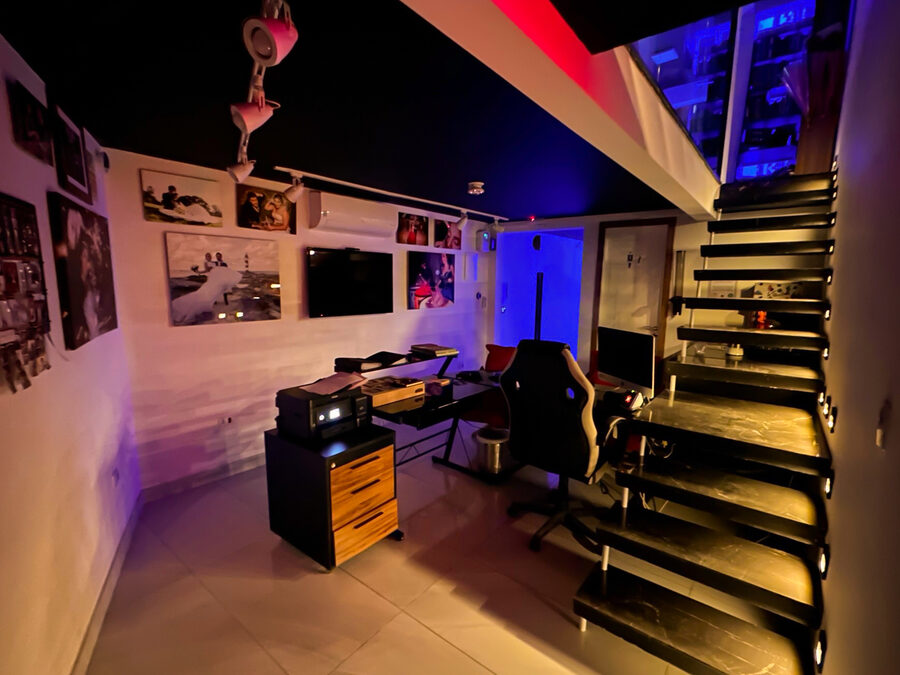 Lounge interno com escada iluminada, quadros na parede e bancada de trabalho