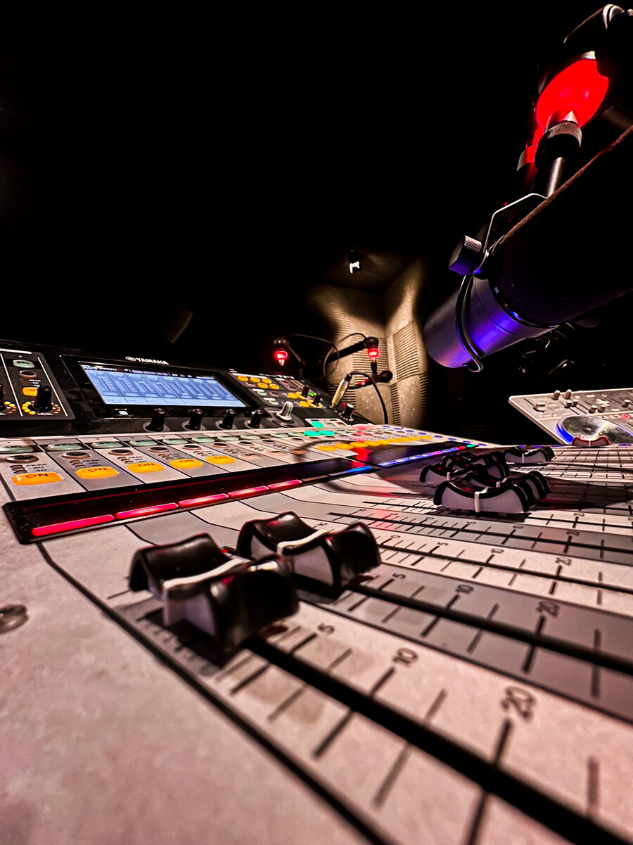 Detalhe rasante dos faders e knobs da mesa de som, com microfone ao fundo