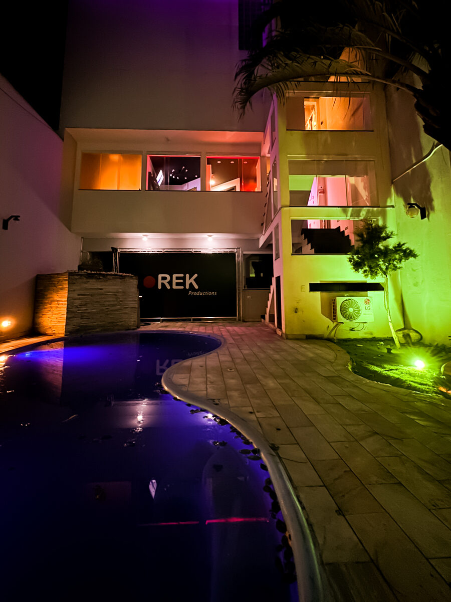 Área externa da REK à noite: piscina iluminada em roxo e verde diante da fachada com a marca REK